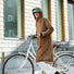 Femme portant la veste imperméable Imbris camel, capuche relevée et vélo à la main lors d’un trajet urbain.