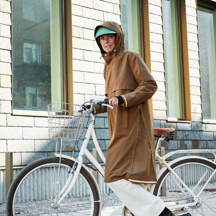 Femme portant la veste imperméable Imbris camel, capuche relevée et vélo à la main lors d’un trajet urbain.