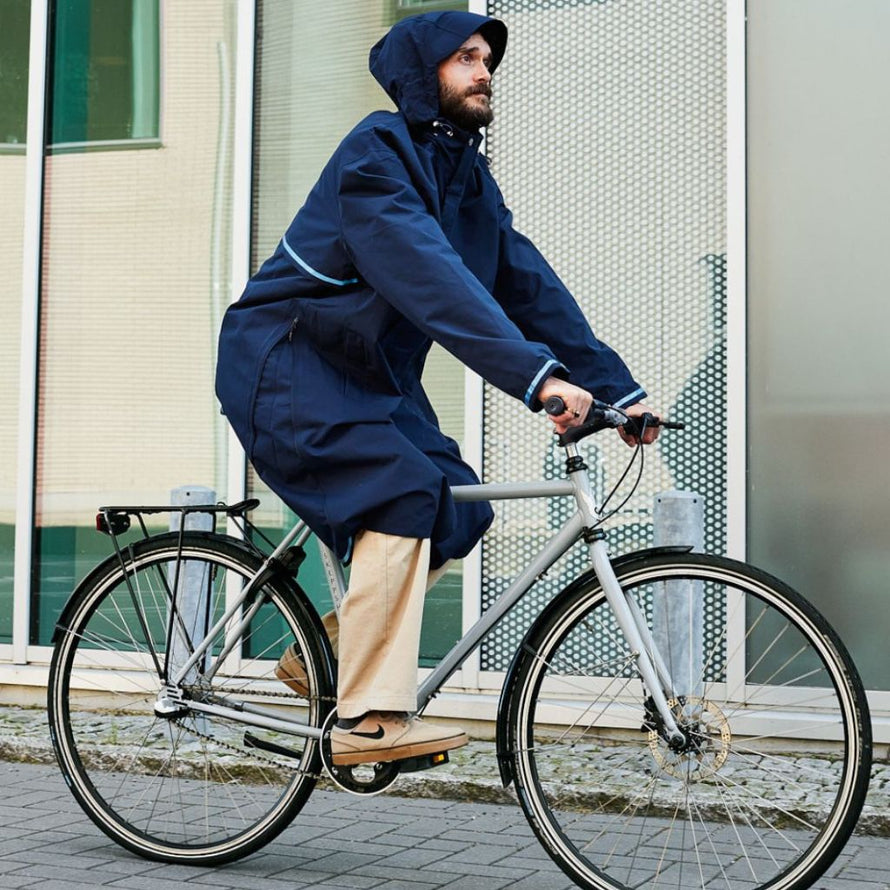 Homme en déplacement à vélo avec la veste imperméable Imbris marine, capuche relevée et détails réfléchissants visibles.