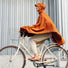 Femme sur un vélo avec le poncho cuivre Imbris Weathergoods Sweden, couvrant efficacement les jambes et protégeant de la pluie.