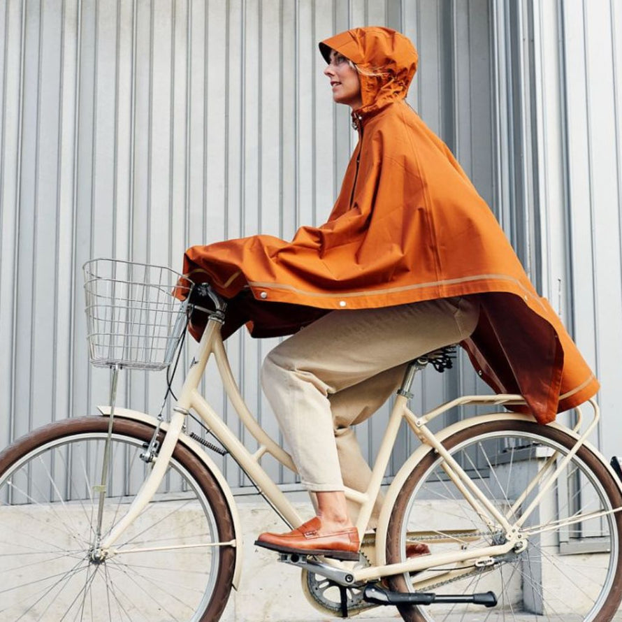 Femme sur un vélo avec le poncho cuivre Imbris Weathergoods Sweden, couvrant efficacement les jambes et protégeant de la pluie.