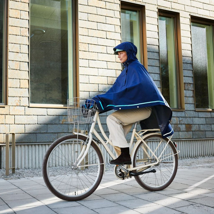 Femme à vélo portant le poncho bleu Imbris Weathergoods Sweden, capuche ajustée et bande réfléchissante ton sur ton.